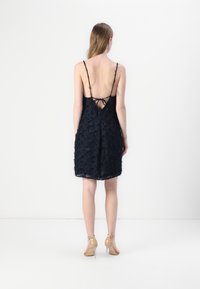 Robe en dentelle noire avec des bretelles spaghetti ajustables, un dos basse avec un système de nouage, et un motif floral texturé. Talons transparents avec bride à la cheville visibles.