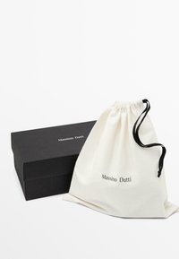 Bawełniany worek sznurkowy w kolorze off-white z czarnym logo, obok czarnego kartonowego pudełka z tą samą nazwą marki wydrukowaną na białym tle.