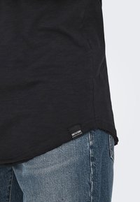 Schwarzes T-Shirt mit Rundhalsausschnitt, aus weichem Stoff; verfügt über ein Logo-Tag am Saum und wird mit blauen verwaschenen Jeans kombiniert.