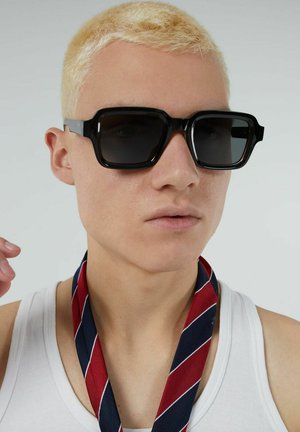 Zwarte oversized zonnebril met een dik montuur, donkere glazen en transparante accenten aan de zijkanten. De drager is bloot bovenlijf met een gestreepte halsaccessoire in rood en blauw.