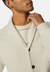 Cremfarbener Kardigan aus geripptem Strickmaterial. Verfügt über einen Schalkragen und drei dunkle Knöpfe, getragen über einem hellbeigen gestreiften Hemd.
