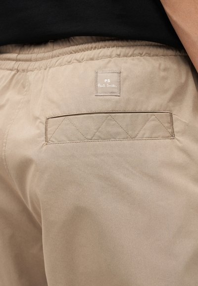 Μπεζ παντελόνια με ελαστική μέση, ραμμένη πίσω τσέπη και ραμμένη τετράγωνη ετικέτα με την επιγραφή "PS Paul Smith" στο πάνω μέρος της πλάτης.