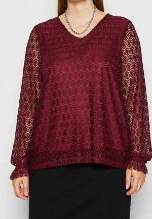 Vrouw draagt een bordeauxrode kanten blouse met lange mouwen met cirkelvormige patronen en een zilveren gedraaide ketting.