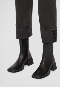 Svarta läderankelboots med fyrkantig tå och klack i blockstil. Har en dragkedja på sidan och en texturerad yta, kombinerat med uppvikta mörka jeans.