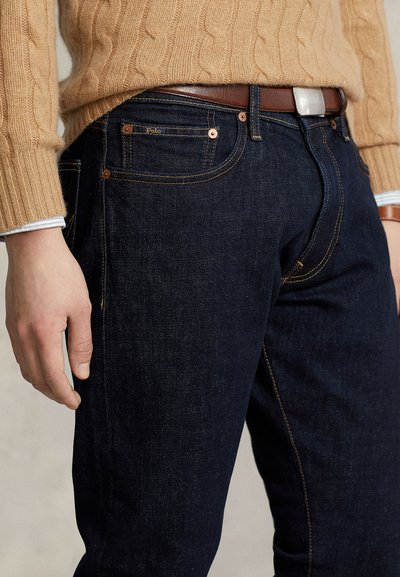 Hombre vistiendo vaqueros azul oscuro con cinturón marrón y suéter beige de punto trenzado, mano relajada a un lado, etiqueta visible de la marca Polo en el bolsillo.