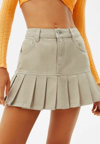 Beige pleitet denimhame med høj talje, frontlommer og knaplukning. Teksturen er glat med en struktureret design.