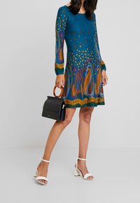 Femme portant une robe bleue à motifs floraux avec des manches longues, tenant un sac à main noir avec une poignée circulaire marron, et portant des talons blancs.