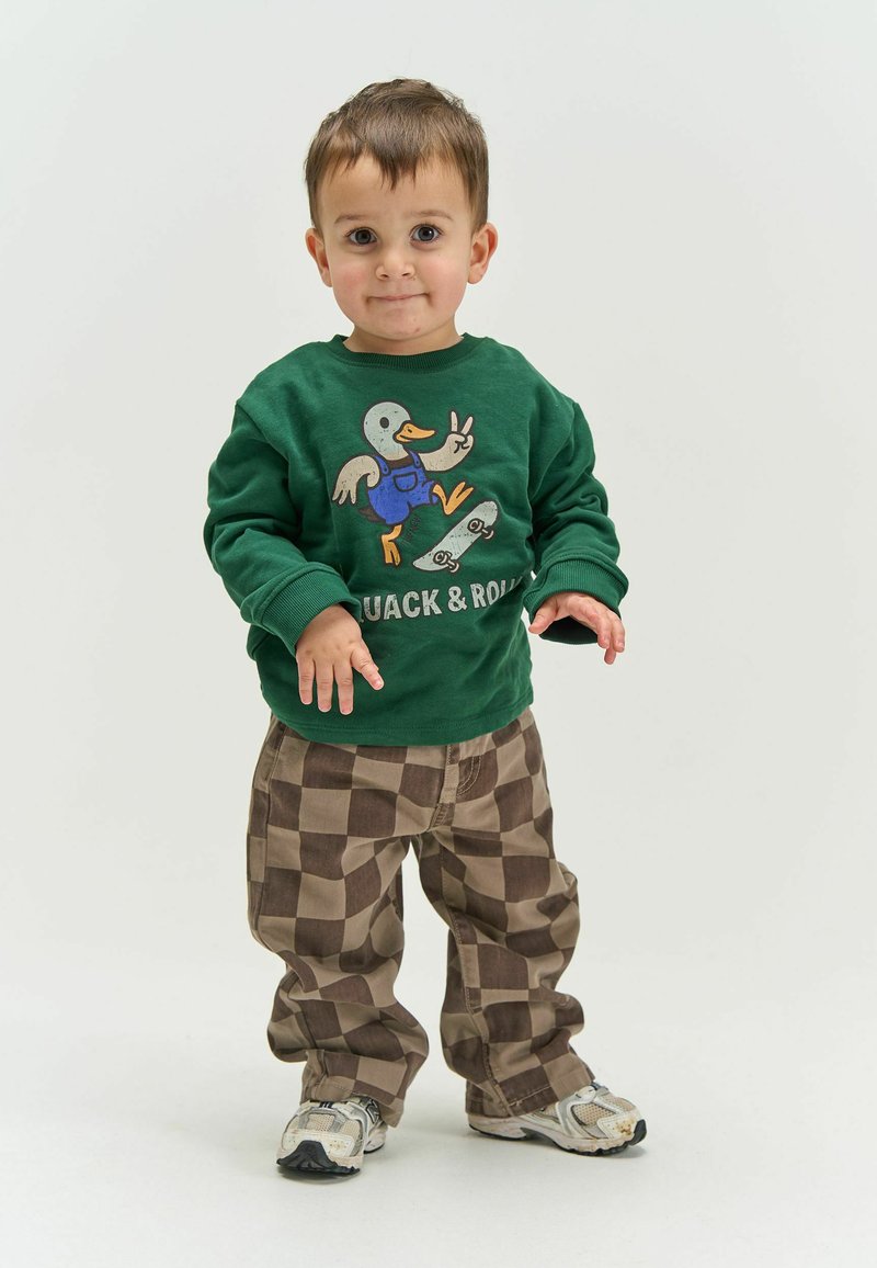 Jeune enfant debout, portant un sweat-shirt vert avec un motif de canard en skate et le texte « Quack & Roll », un pantalon marron à carreaux et des baskets.
