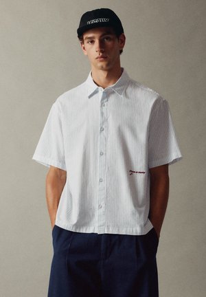 Jeune homme portant une chemise blanche à manches courtes avec de fines rayures et un pantalon bleu marine, debout les mains dans les poches et une casquette noire.