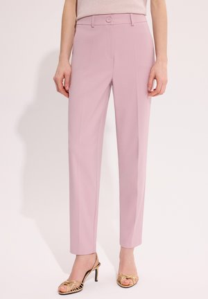 Kobieta ubrana w różowe spodnie z wysokim stanem o prostych nogawkach, w zestawieniu ze złotymi sandałami na wysokim obcasie z odkrytymi palcami, stojąca na jasnym tle.