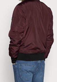 Chaqueta bomber burdeos con una textura lisa y brillante, puños y dobladillo acanalados, bolsillo lateral con cremallera y corte relajado. Lleva puesta sobre unos vaqueros azules.