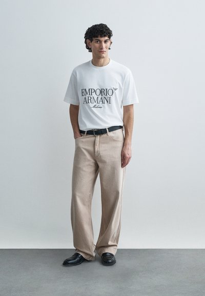 Biele tričko s krátkym rukávom s čiernym nápisom "EMPORIO ARMANI", skombinované s béžovými širokými nohavicami a čiernymi topánkami. Jednoduchý, moderný dizajn.