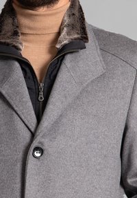 Abrigo gris con cuello de piel, tejido texturizado, capa interior negra con cremallera y cierre de botón. Detalle de diseño suave y estructurado visible.