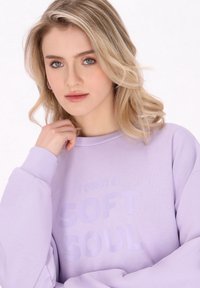 Lila mikina s kulatým výstřihem a dlouhými rukávy, s embosovaným textem "Mám MĚKKOU DUŠI." Měkká látka s texturovaným povrchem.