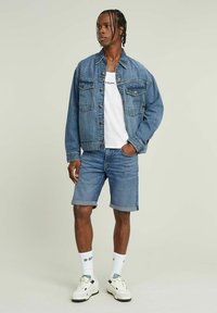 Veste en denim avec fermeture à boutons et poches poitrine, associée à un short en denim retroussé. T-shirt blanc et baskets blanches, avec un imprimé logo.