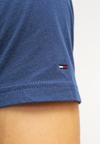 Modrá bavlněná tričko s krátkými rukávy a šitým lemem, na rukávu je malé vyšívané logo v červené, bílé a modré barvě.