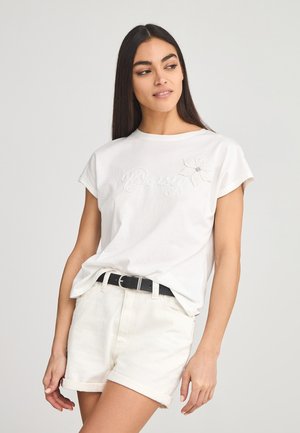 T-shirt bianco in cotone con maniche corte, caratterizzato da testo ricamato "Daisy" e un design floreale. Abbinato a pantaloni corti in denim di colore chiaro.
