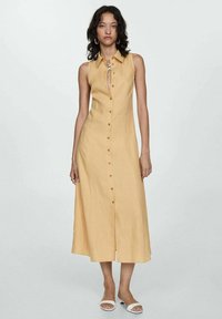 Donna con capelli scuri e ondulati, che indossa un vestito midi beige senza maniche con bottoni e sandali bianchi aperti, in piedi su uno sfondo chiaro e uniforme.