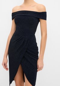 Robe midi noire texturée à épaules dénudées avec un corsage ajusté et une fente asymétrique devant, portée par une personne sur un fond uni.