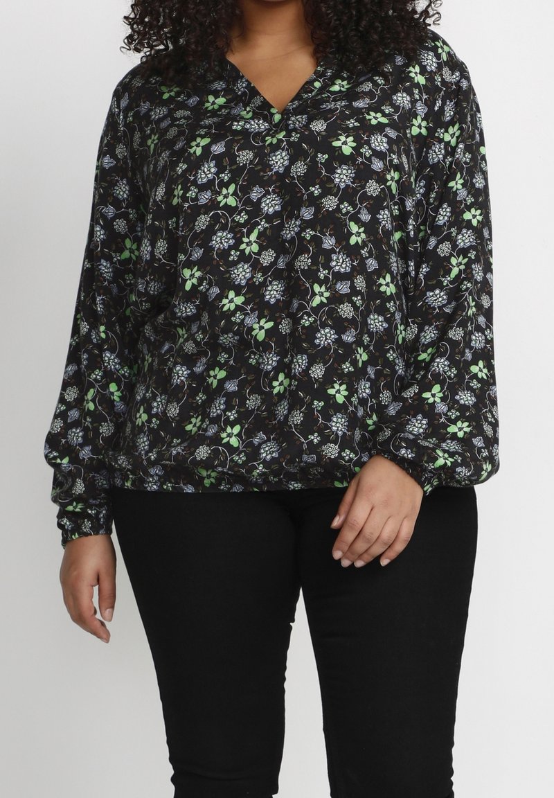 Blouse fleurie noire à manches longues, avec des motifs verts et bleus, col en V et poignets élastiques. Associée à un pantalon noir.