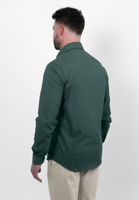 Donkergroen shirt met een button-down kraag en lange mouwen. Heeft een zachte textuur en een rechte zoom. Draagt met beige broeken.