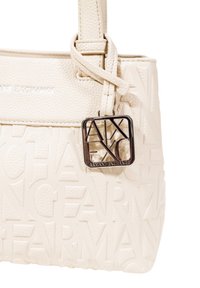 Borsa di colore crema con logo del brand in rilievo, pelle texture e un ciondolo con logo in tonalità argento su una tracolla decorativa.