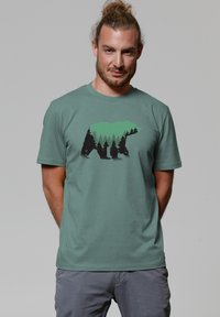 T-shirt vert menthe avec un graphique d'un ours et des arbres dans des tones noirs et verts. Manches courtes et encolure ras du cou. Texture de tissu douce.