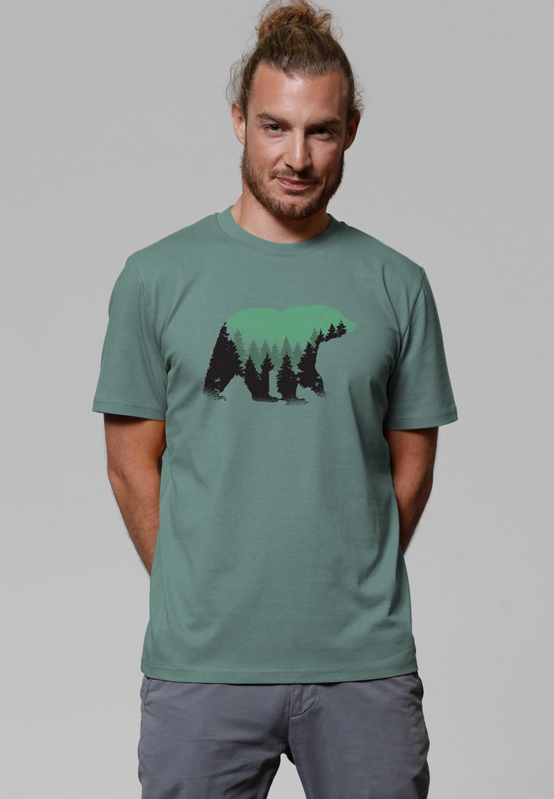 T-shirt vert menthe avec un graphique d'un ours et des arbres dans des tones noirs et verts. Manches courtes et encolure ras du cou. Texture de tissu douce.
