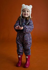 Kombinezon w kwiatowy wzór w kolorze fioletowym z czarnymi akcentami, noszony z różowymi gumowymi butami i kremową czapką z uszami misia, dziecko uśmiechające się, z rękami wyciągniętymi.