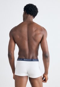 Boxers blancos ajustados con una cintura azul marino que presenta el nombre "GANT" impreso varias veces. Textura suave, diseño ceñido, vista trasera de la prenda.