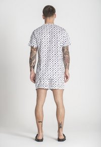 El pijama blanco de manga corta presenta un patrón repetido con logotipos negros, tejido suave y pantalones cortos hasta la rodilla con una cinturilla elástica.