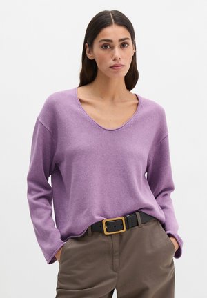 Femme portant un pull à manches longues lilas rentré dans un pantalon marron avec une ceinture noire, debout les mains dans les poches devant un fond uni.