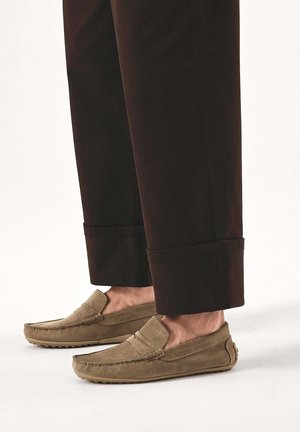 Braune Wildlederloafers getragen zu schwarzen, umgeschlagenen Hosen vor hellem Hintergrund, mit seitlichen und oberen Details der Schuhe.