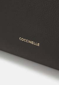 Mustaa teksturoitua nahkaa, jossa on kultapainettu "COCCINELLE"-logo. Pinta esittää hienoa urapintakuviota materiaalin yli.