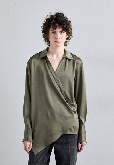 Chemise à manches longues vert olive en matériau satiné avec un design drapé, encolure en V et ourlet asymétrique, associée à un jean foncé.