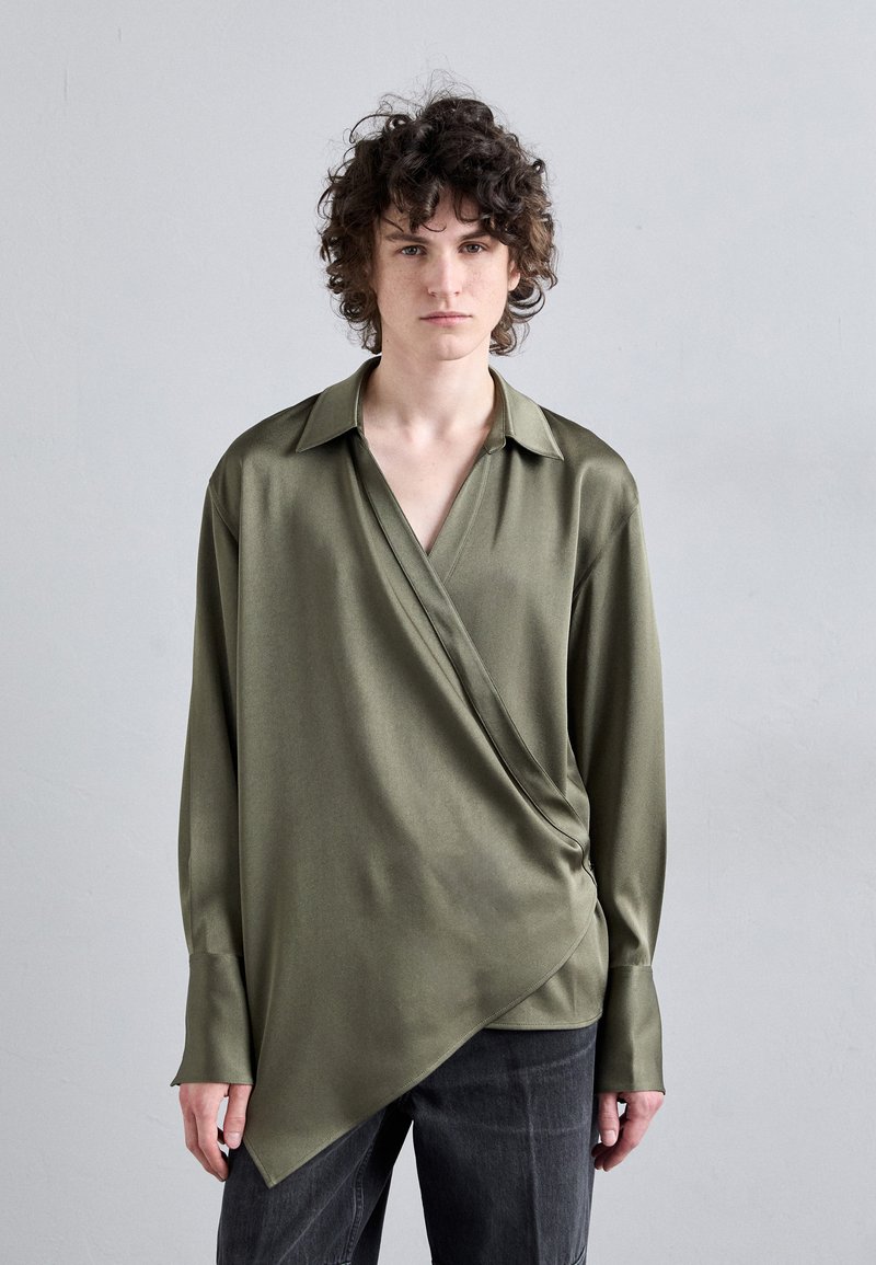 Olivgrünes Langarmhemd aus satinähnlichem Material mit Wickeldesign, V-Ausschnitt und asymmetrischem Saum, kombiniert mit dunklen Jeans.