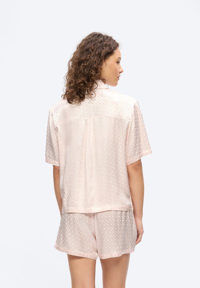 Femme aux cheveux bouclés portant un t-shirt à manches courtes rose pâle et un short assorti en tissu à motif subtil, vue de dos sur fond blanc.