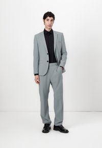 Lichtgrijze colbert met bijpassende broek, slanke pasvorm, gestructureerde stof, zwart overhemd eronder en glanzende zwarte schoenen.