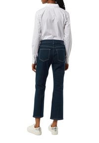 Dunkelblaue, verkürzte Jeans mit kontrastierenden Nähten, fünf Taschen und einer taillierten Silhouette, kombiniert mit einem weißen Hemd und weißen Sneakers.