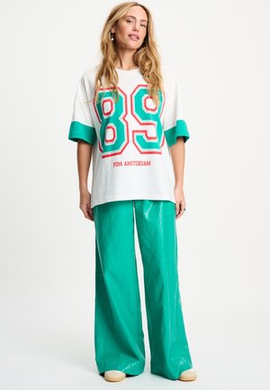 Vrouw met lang blond haar die een wit oversized T-shirt draagt met het groene en rode nummer 89 en turquoise wijde broek, staand op een witte achtergrond.
