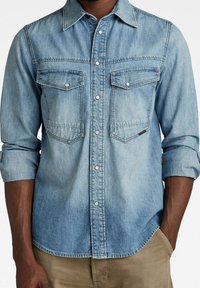 Lichtblauwe denim overhemd, voorzien van twee borstzakken, drukknoopjes en een verspreide kraag. Lange mouwen opgerold bij de manchetten.