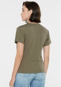 Olive green cotton T-shirt with short sleeves, a round neck, and a smooth texture. Side stitching detail visible; worn with light blue denim.