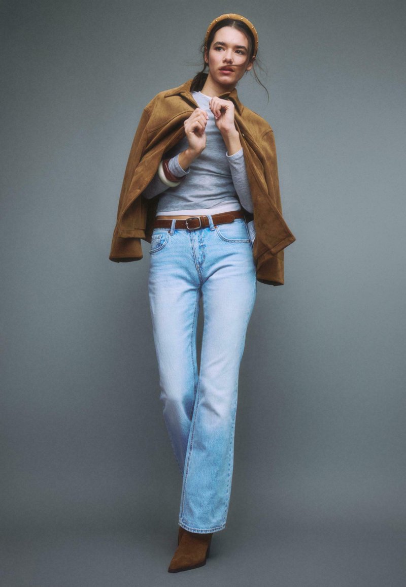 Donna con giacca marrone sulle spalle, top grigio, jeans azzurri, cintura marrone, stivali marroni e fascia gialla su sfondo grigio.