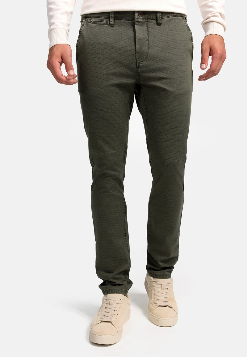 Oliwkowe, slim-fit chinos wykonane z gładkiego materiału, z delikatnym wzorem strukturalnym i standardowymi kieszeniami. Noszone z jasnymi trampkami.
