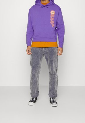 Hoodie violet avec impressions de logo, chemise orange superposée en dessous, pantalon en velours côtelé gris, et baskets noires avec lacets blancs.