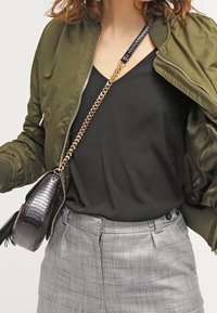 Olivově zelená bomber bunda, černý top, šedé kostkované kalhoty a malá černá taška s řetízkovým popruhem a texturovanými detaily.