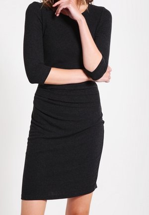 Kvinde iført en tætsiddende sort knælange kjole med trekvartlange ærmer, stående med armene krydset og hånden nær ansigtet.