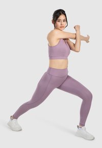 Sostenedor deportivo de color púrpura claro y leggings de cintura alta usados por un modelo en una pose de estiramiento dinámico. Zapatillas deportivas blancas, textura suave, diseño ajustado.