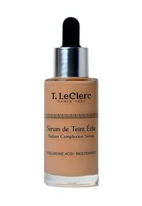Strahlendes Teint-Serum in einer Glasflasche. Beigefarbene Flüssigkeit im Inneren mit einem schwarzen Tropfer und silberner Kappe. Der Text enthält Marke und Produktdetails.