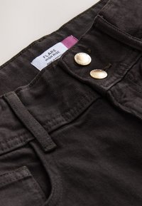 Gros plan sur un jean flare taille haute noir avec double boutons métalliques et une étiquette blanche indiquant « FLARE HIGH RISE Made in Pakistan. »